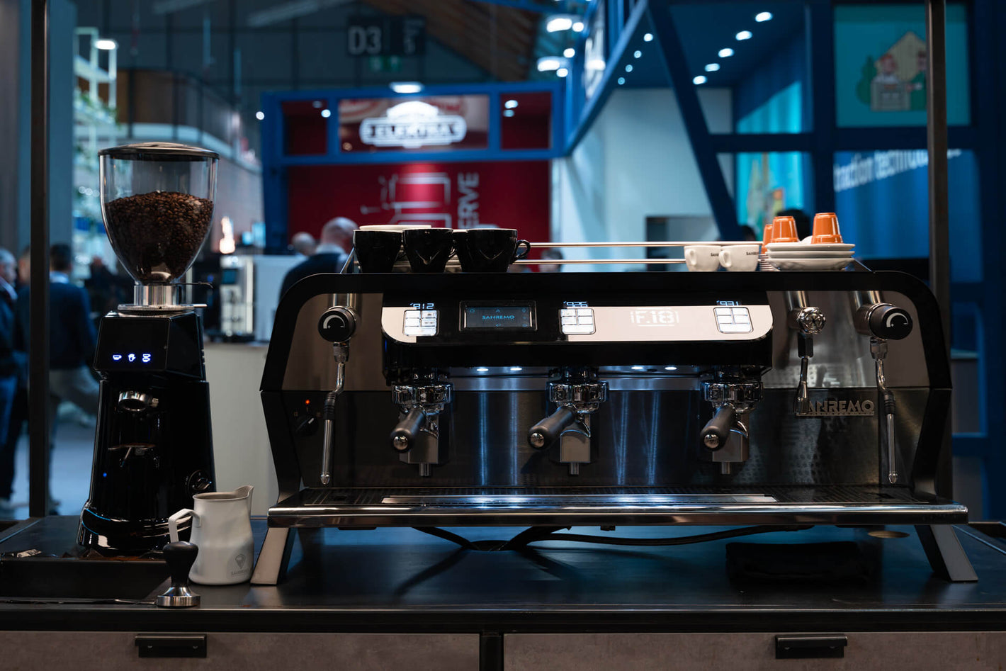 Sanremo F18 3 groups black commercial espresso machine and coffee grinder front view, at a coffee industry event